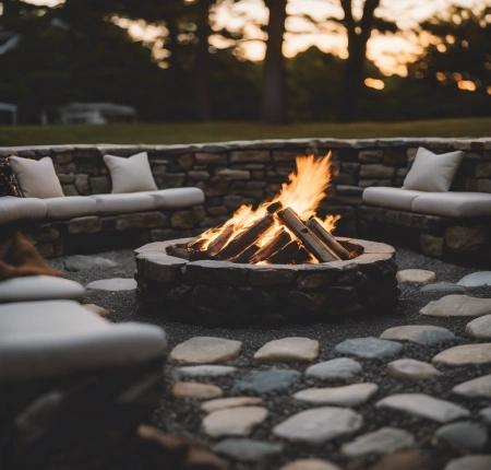 Rustic stone fire pit with a wood-burning fire in a backyard garden, surrounded by blooming flowers and green foliage.
